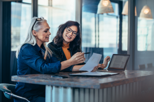 two women looking at a piece of paper in front of a laptop computer.