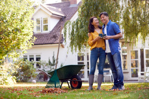 a man and a woman holding blue coffee cups together while raking leaves in a grass covered yard in front of a white house with trees.